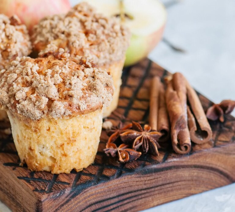 Drei Apfel-Streusel-Muffins auf einem Brett mit Anissternen und Zimtstangen, im Hintergrund unscharf halbierte Äpfel. | © Adobe Stock/ galiyahassan