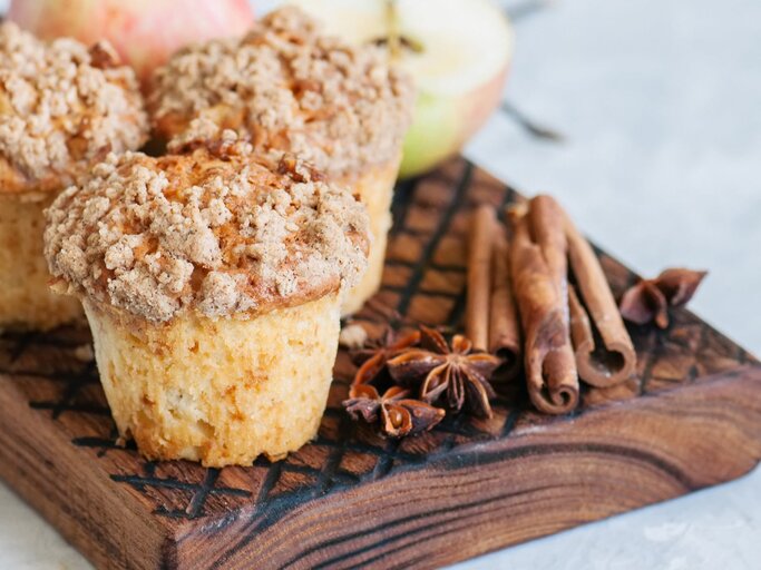 Drei Apfel-Streusel-Muffins auf einem Brett mit Anissternen und Zimtstangen, im Hintergrund unscharf halbierte Äpfel. | © Adobe Stock/ galiyahassan