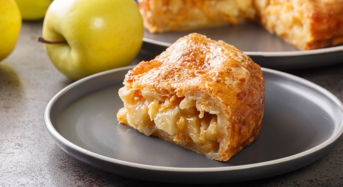 Ein Stück vom Apfel-Cheddar-Kuchen auf einem grauen Teller mit frischen Äpfeln im Hintergrund.