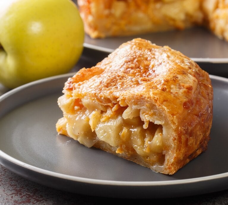 Ein Stück vom Apfel-Cheddar-Kuchen auf einem grauen Teller mit frischen Äpfeln im Hintergrund. | © Shutterstock/ ALLEKO