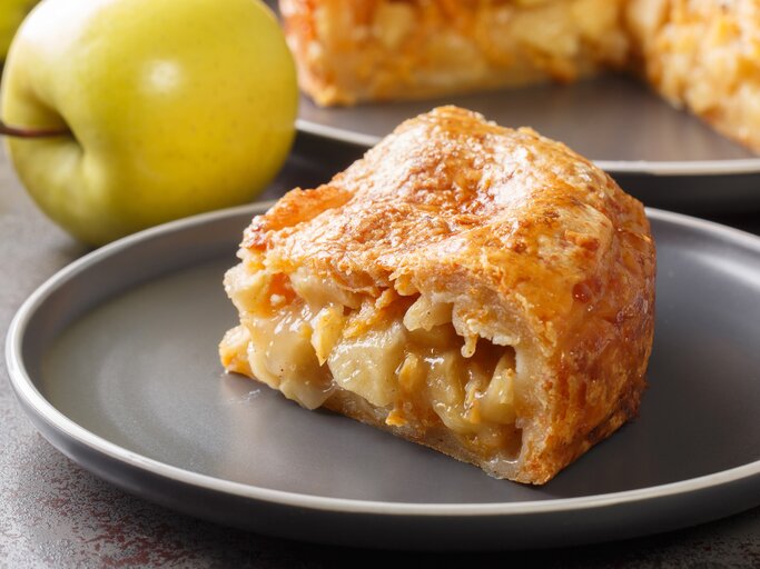 Ein Stück vom Apfel-Cheddar-Kuchen auf einem grauen Teller mit frischen Äpfeln im Hintergrund. | © Shutterstock/ ALLEKO