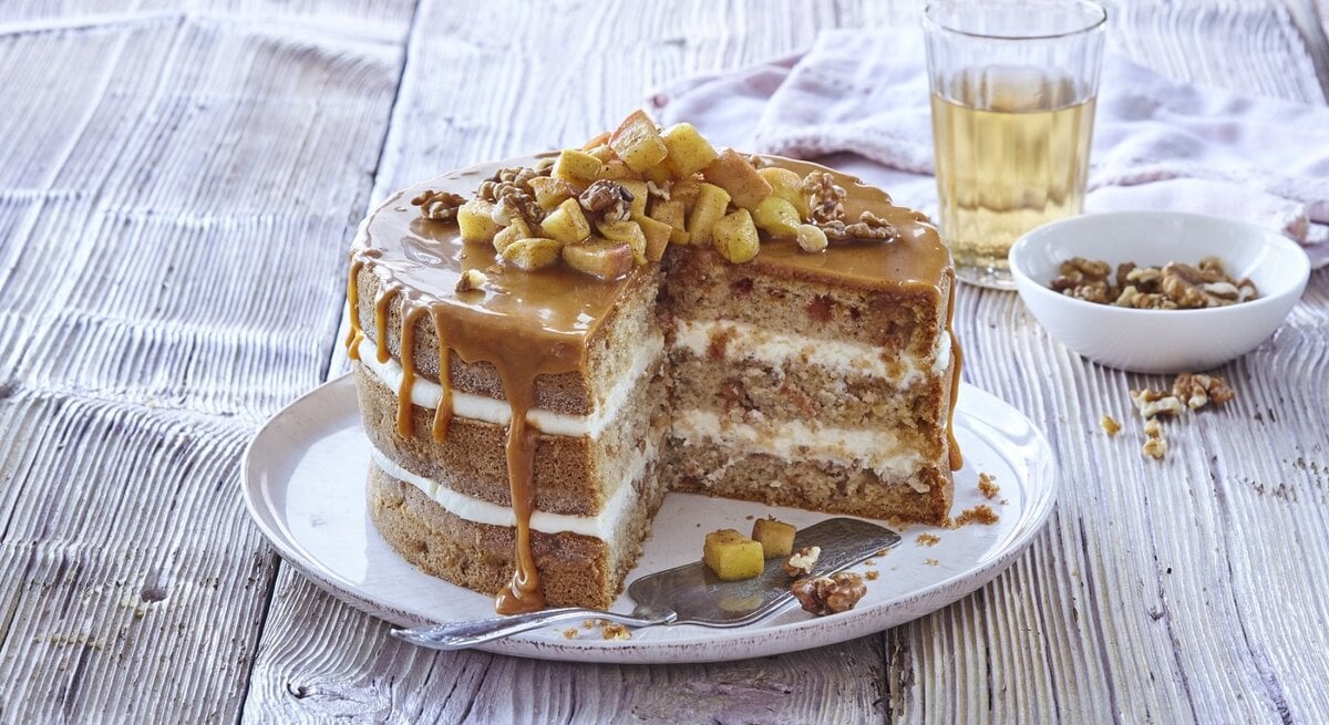 Auf einem Teller steht eine Apfel-Karamell-Schichttorte. Sie ist mit Äpfeln und Nüssen bedeckt. 