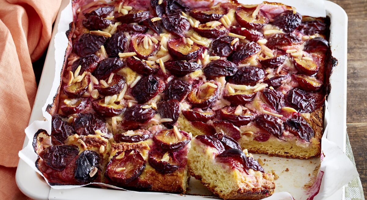 Angeschnittener Marzipan-Pflaumenkuchen in weißem Blech auf einem karierten Küchentuch, darunter Holz. 