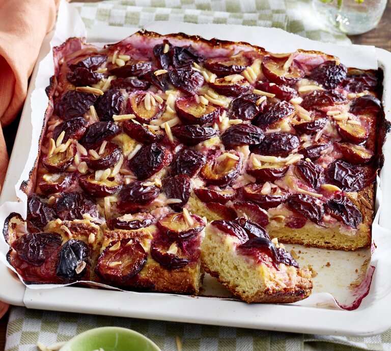 Angeschnittener Marzipan-Pflaumenkuchen in weißem Blech auf einem karierten Küchentuch, darunter Holz.  | © EAT CLUB / Isabell Triemer 