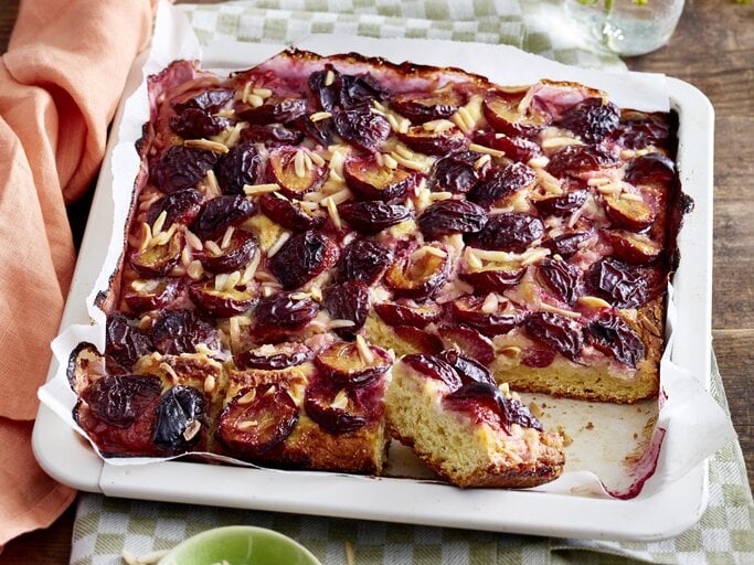 Angeschnittener Marzipan-Pflaumenkuchen in weißem Blech auf einem karierten Küchentuch, darunter Holz.  | © EAT CLUB / Isabell Triemer 