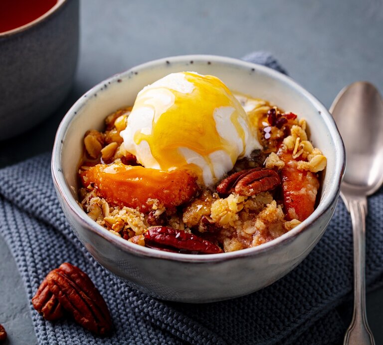 Eine Schale mit Pfirsich-Crumble auf einem grauen Küchentuch. | © Anna_Pustynnikova