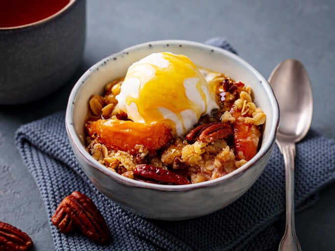 Eine Schale mit Pfirsich-Crumble auf einem grauen Küchentuch. | © Anna_Pustynnikova