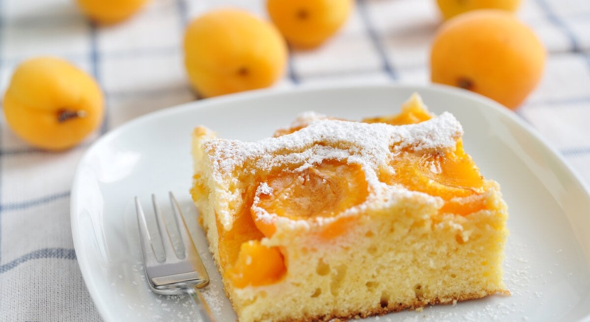 Ein Teller mit einem Stück fluffiger Aprikosen-Marzipan-Kuchen steht auf einen Handtuch. Daneeben liegen viele Aprikosen, beziehungsweise marillen.