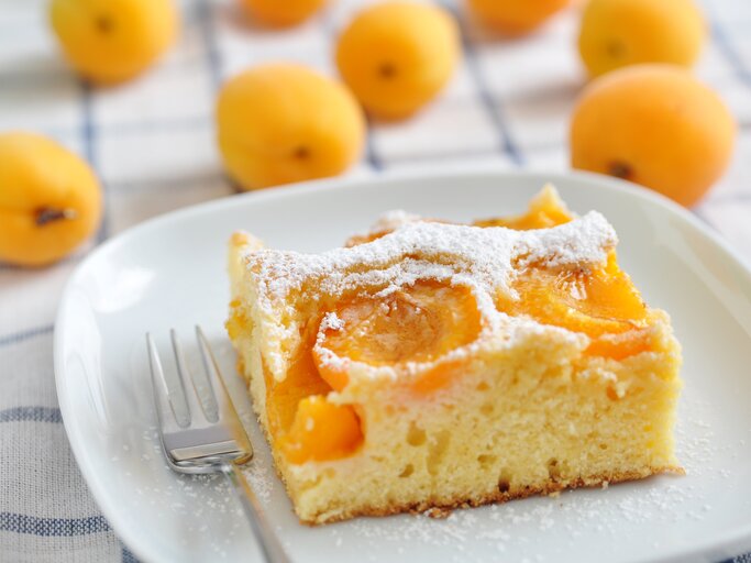 Ein Teller mit einem Stück fluffiger Aprikosen-Marzipan-Kuchen steht auf einen Handtuch. Daneeben liegen viele Aprikosen, beziehungsweise marillen. | © Shutterstock / A_Lein