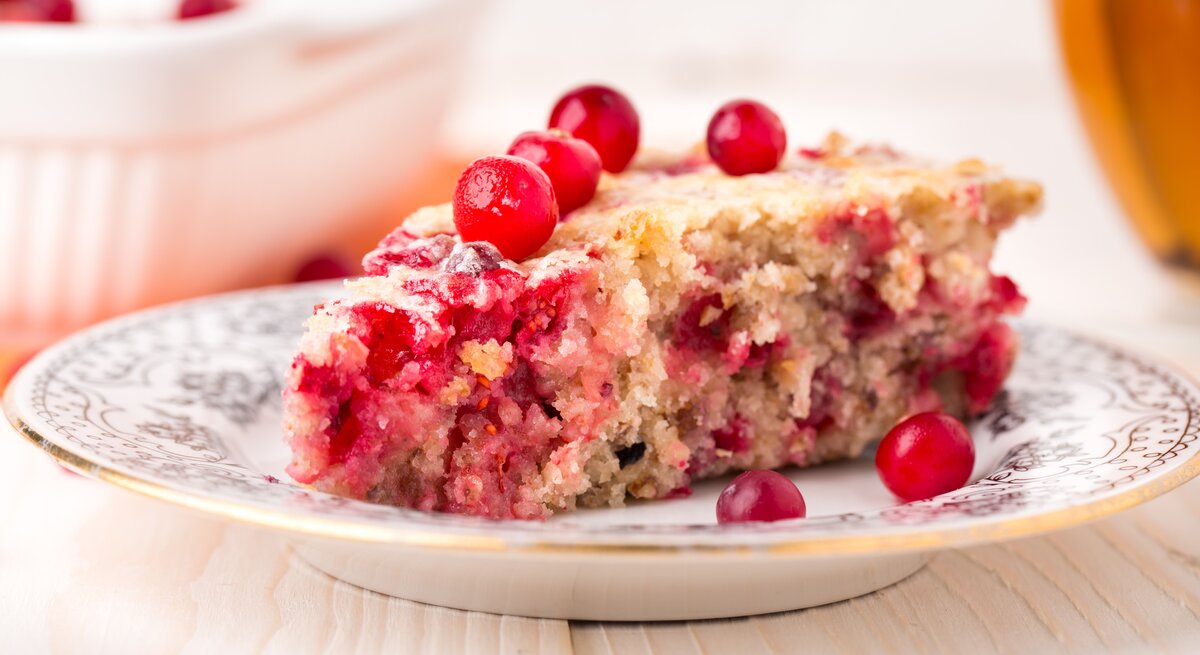 Ein weißer Teller mit Verzierungen und einem Stück vom Johannisbeerkuchen auf einem hellen Holztisch seitlich fotografiert.