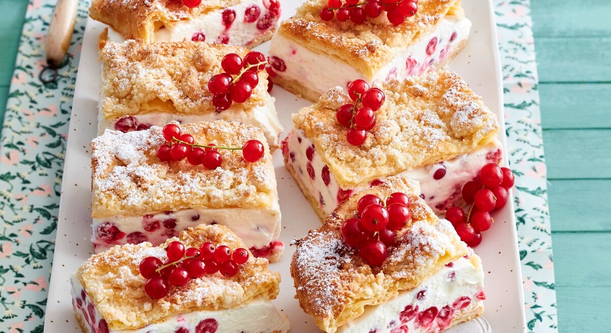 Sechs viereckige Johannisbeer-Knusperschnitten mit frischen Johannisbeeren auf einem rosa Teller auf einem gemusterten Tuch mit blauem Holzuntergrund. 