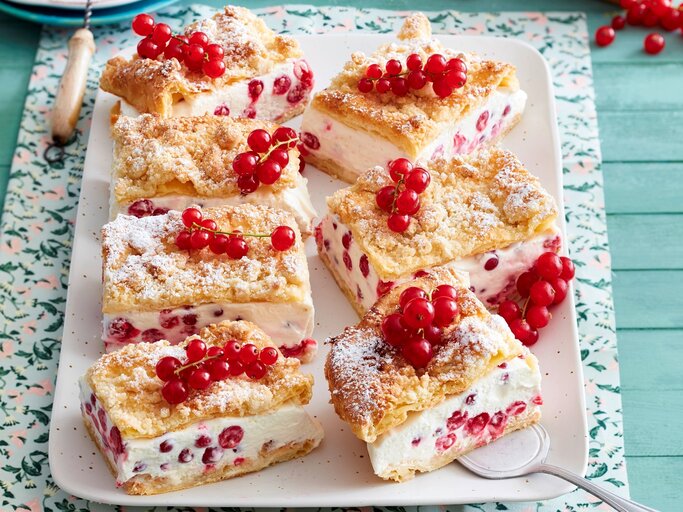 Sechs viereckige Johannisbeer-Knusperschnitten mit frischen Johannisbeeren auf einem rosa Teller auf einem gemusterten Tuch mit blauem Holzuntergrund.  | © EAT CLUB / Isabell Triemer