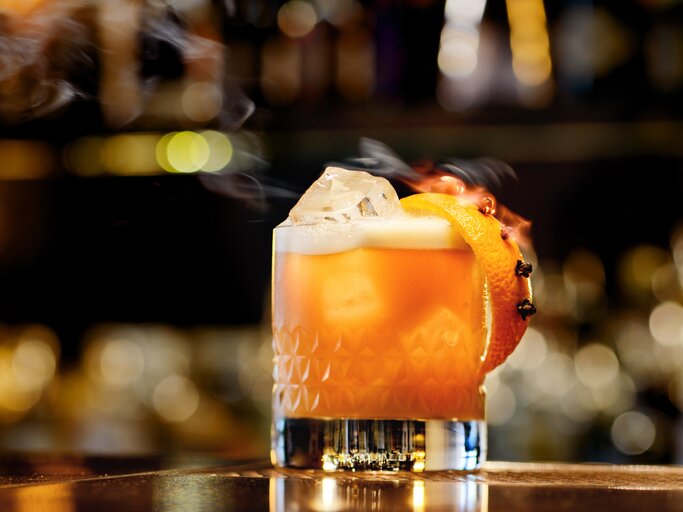 Ein Glas Whisky Sour steht auf einem Bartresen. Im Hintergrund verschwimmen einige Lichter der Bar. Das Glas ist angerichtet mit einer Orangenscheibe. Im Cocktail schwimmt ein Eiswürfel. | © Getty Images / Jordan Lye