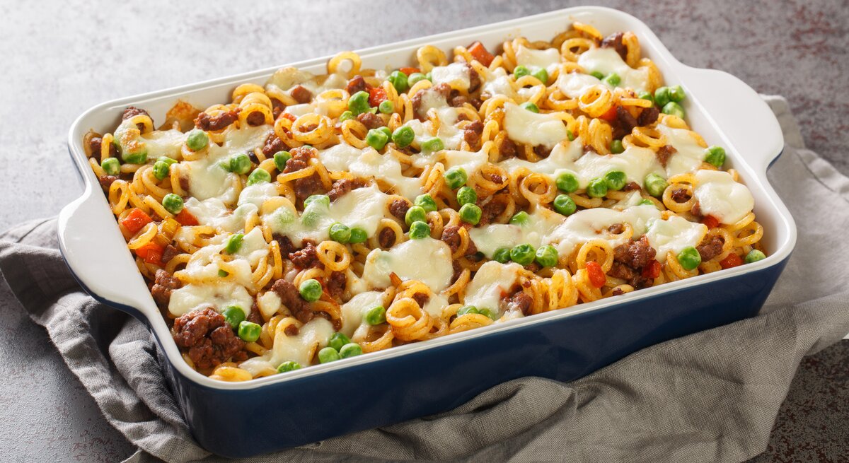Ein appetitanregender Nudelauflauf in einer blauen Keramikform, reichlich bestreut mit grünen Erbsen und geschmolzenem Käse. Die Pasta scheint mit einer Fleischsauce vermengt zu sein, und das Ganze präsentiert sich als herzhaftes, überbackenes Gericht, das auf einem grauen Stofftuch ruht und auf einer strukturierten Arbeitsfläche angerichtet ist.