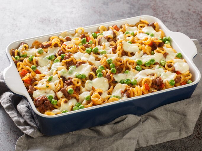 Ein appetitanregender Nudelauflauf in einer blauen Keramikform, reichlich bestreut mit grünen Erbsen und geschmolzenem Käse. Die Pasta scheint mit einer Fleischsauce vermengt zu sein, und das Ganze präsentiert sich als herzhaftes, überbackenes Gericht, das auf einem grauen Stofftuch ruht und auf einer strukturierten Arbeitsfläche angerichtet ist. | © Getty Images/ALLEKO