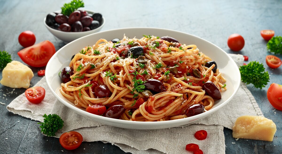 Ein weißer Pastateller mit den Spaghetti alla Puttanesca auf grauem Untergrund mit weißem Leinentuch. Daneben diverse Pastazutaten wie Parmesan, Tomaten, Oliven, Chili und Petersilie.