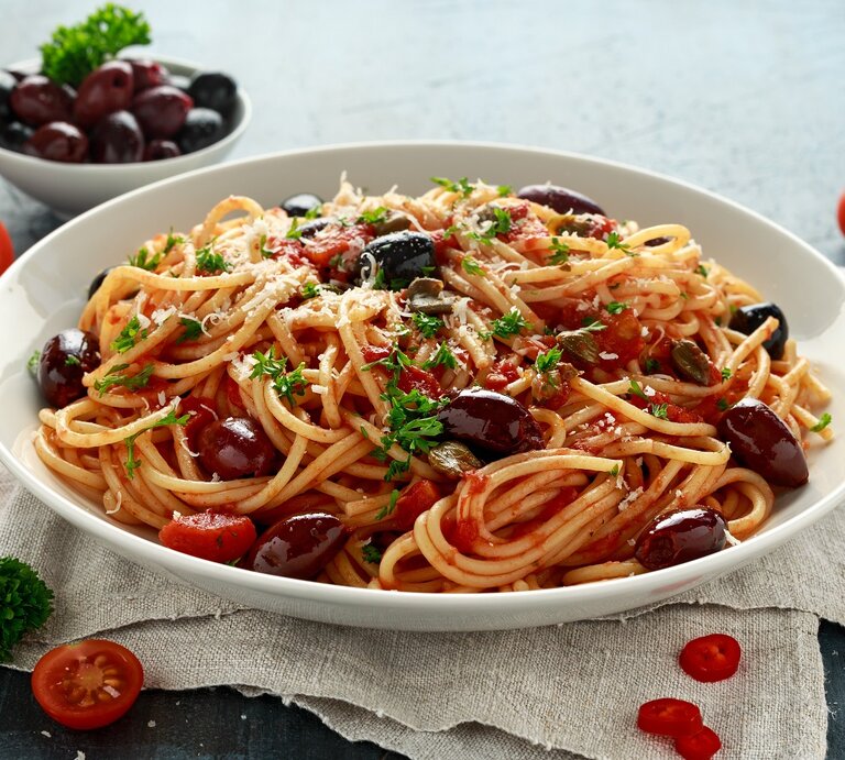 Ein weißer Pastateller mit den Spaghetti alla Puttanesca auf grauem Untergrund mit weißem Leinentuch. Daneben diverse Pastazutaten wie Parmesan, Tomaten, Oliven, Chili und Petersilie. | © Shutterstock/ DronG