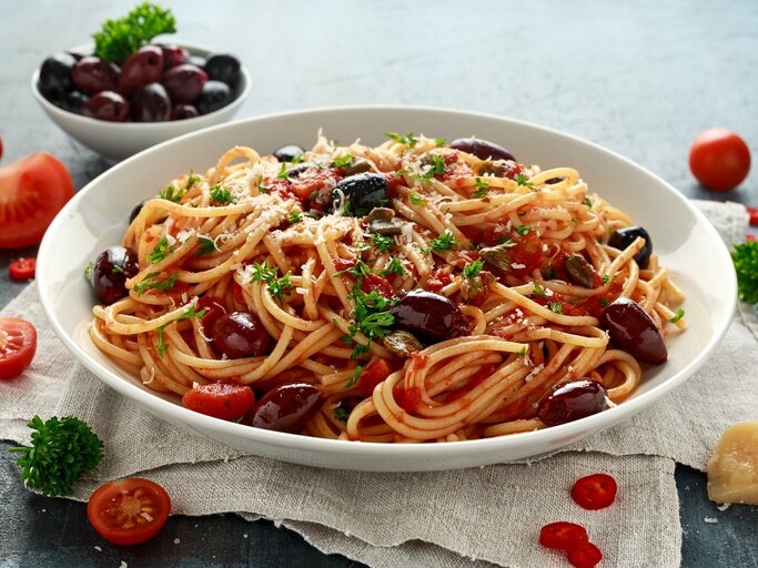 Ein weißer Pastateller mit den Spaghetti alla Puttanesca auf grauem Untergrund mit weißem Leinentuch. Daneben diverse Pastazutaten wie Parmesan, Tomaten, Oliven, Chili und Petersilie. | © Shutterstock/ DronG
