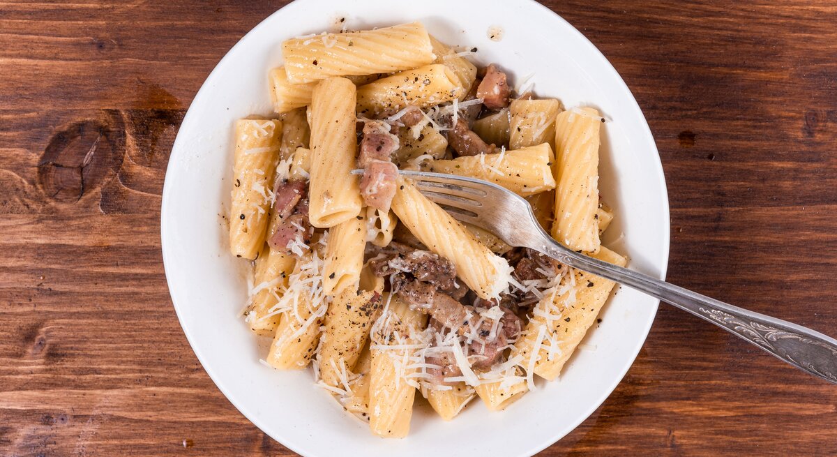 Rigatoni mit gebratenem Guanciale und Pecorino auf weißem Teller. Dieser steht auf einem Holzuntergrund. 