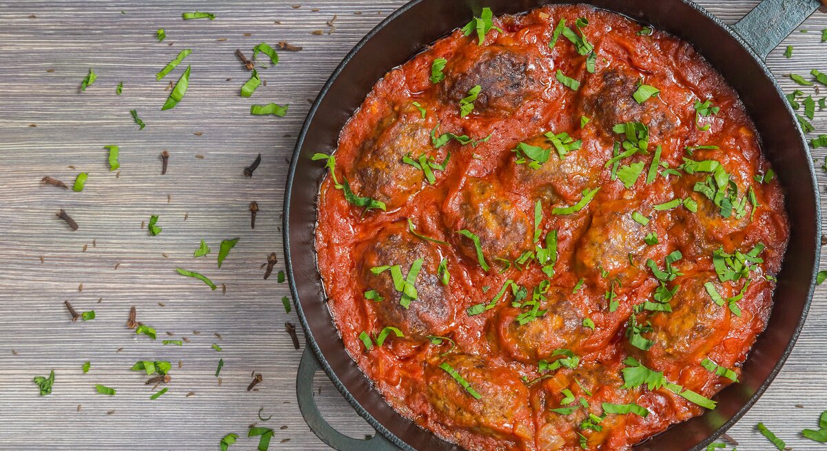 Soutzoukakia in Tomatensoße in schwarzer Pfanne auf Holztisch. 