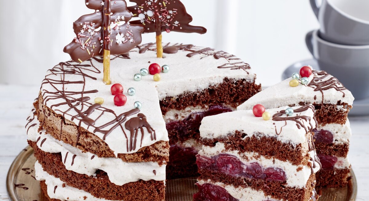 Schwarzwälder Glühwein-Kirschtorte, angeschnitten serviert auf einem Servierteller, dekoriert mit Weihnachtlichem Schmuck wie Lebkuchen und kandierten Kirschen. 