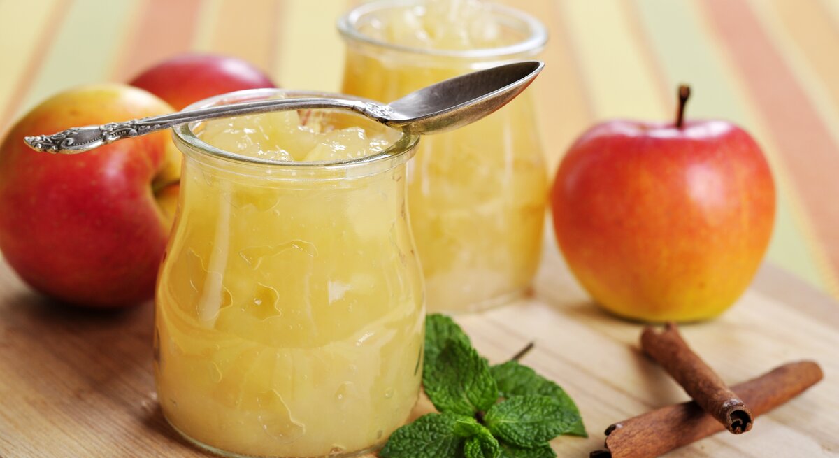 Zwei Gläser Apfel-Minz-Marmelade stehen auf einem Holzbrett. Drumherum liegen Äpfel.