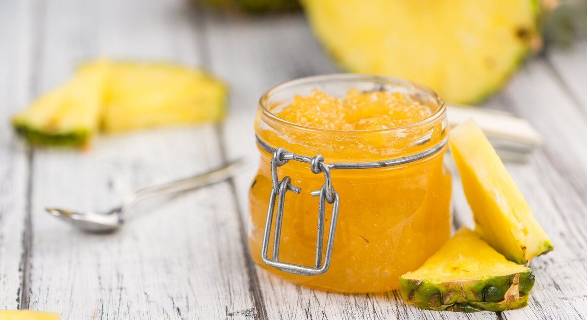 Ein Glas Ananas-Marmelade steht auf einem hellen Dielenboden. Drum herum liegen dreieickige Ananasstücke und ein Löffel. Im Hintergrund eine angeschnittene Ananas.