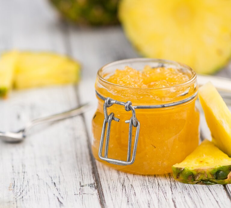 Ein Glas Ananas-Marmelade steht auf einem hellen Dielenboden. Drum herum liegen dreieickige Ananasstücke und ein Löffel. Im Hintergrund eine angeschnittene Ananas. | © Shutterstock / HandmadePictures
