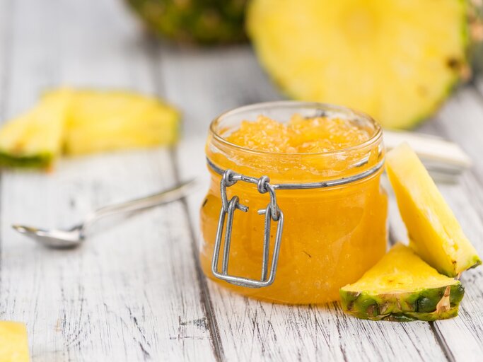 Ein Glas Ananas-Marmelade steht auf einem hellen Dielenboden. Drum herum liegen dreieickige Ananasstücke und ein Löffel. Im Hintergrund eine angeschnittene Ananas. | © Shutterstock / HandmadePictures
