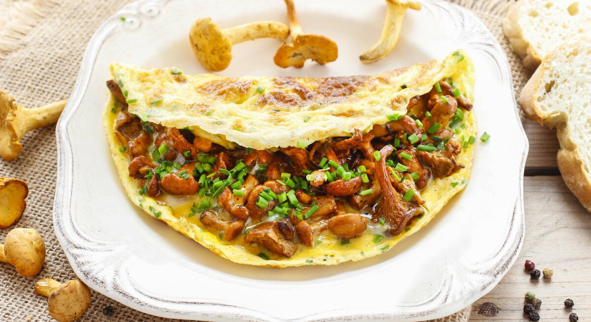 Auf einem weißen Teller liegt ein Pfifferling-Omelette. Um den Teller herum liegen frische Pfifferlinge, Pfefferkörnern und Weißbrotscheiben.