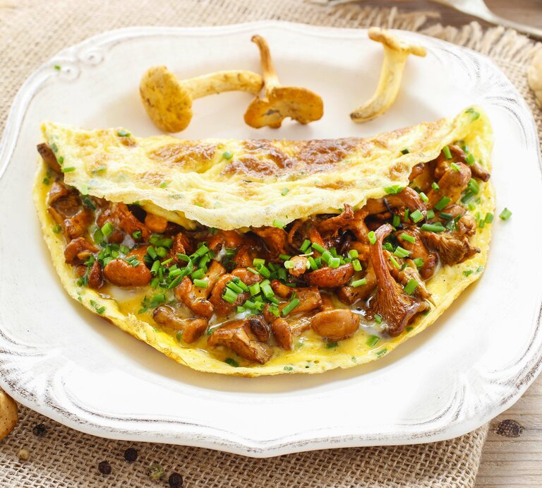 Auf einem weißen Teller liegt ein Pfifferling-Omelette. Um den Teller herum liegen frische Pfifferlinge, Pfefferkörnern und Weißbrotscheiben. | © Shutterstock / Agnes Kantaruk