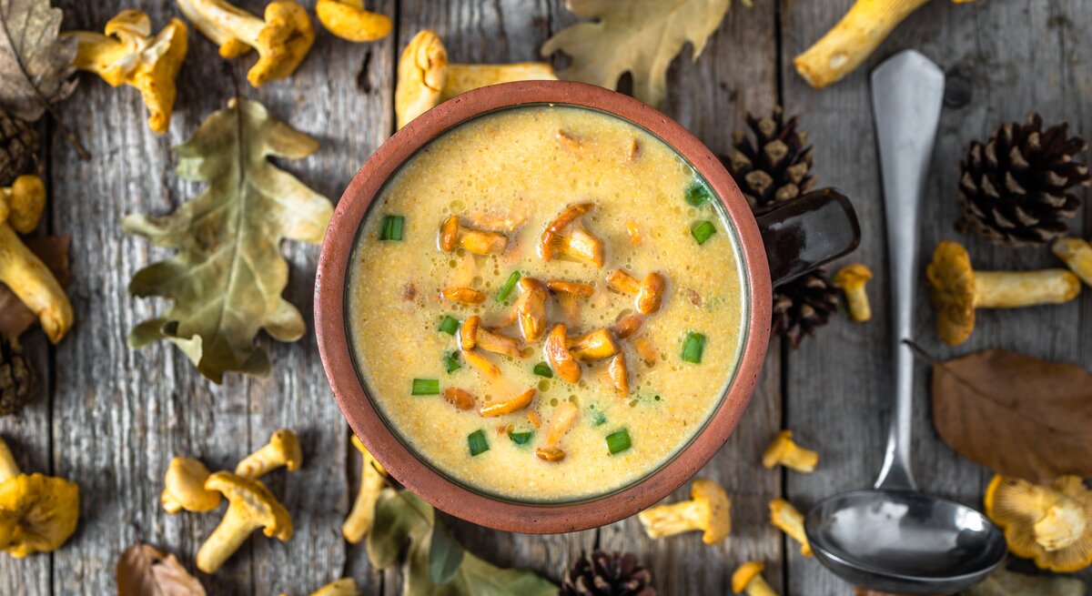 Aus der Vogelperspektive: eine Schüssel Kartoffelsuppe mit Pfifferlingen stehtauf einem Untergund aus Holz. Drumherum liegen Blätter, Tannenzapfen und frische  Pfifferling sowie eine Suppenkelle.