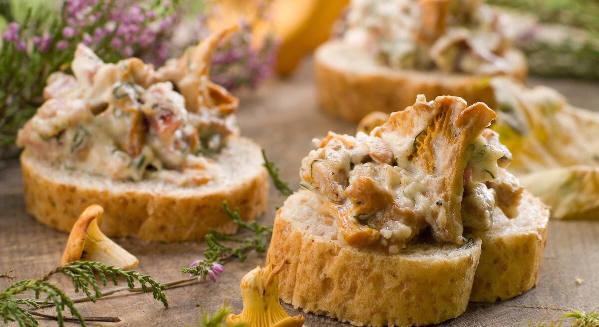 3 Pfifferling-Frischkäse-Toasts auf Baguette, drumherum Blüten und Baumnadeln sowie frische Pfifferlinge.