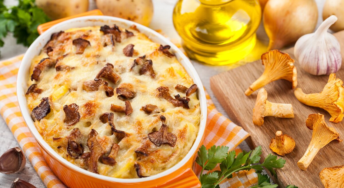 Kartoffelauflauf mit Pfifferlingen in einer gelben Auflaufform, drumherum frische Kartoffeln, Pfifferlinge und Knoblauch, außerdem eine Flasche Öl und Kräuter.