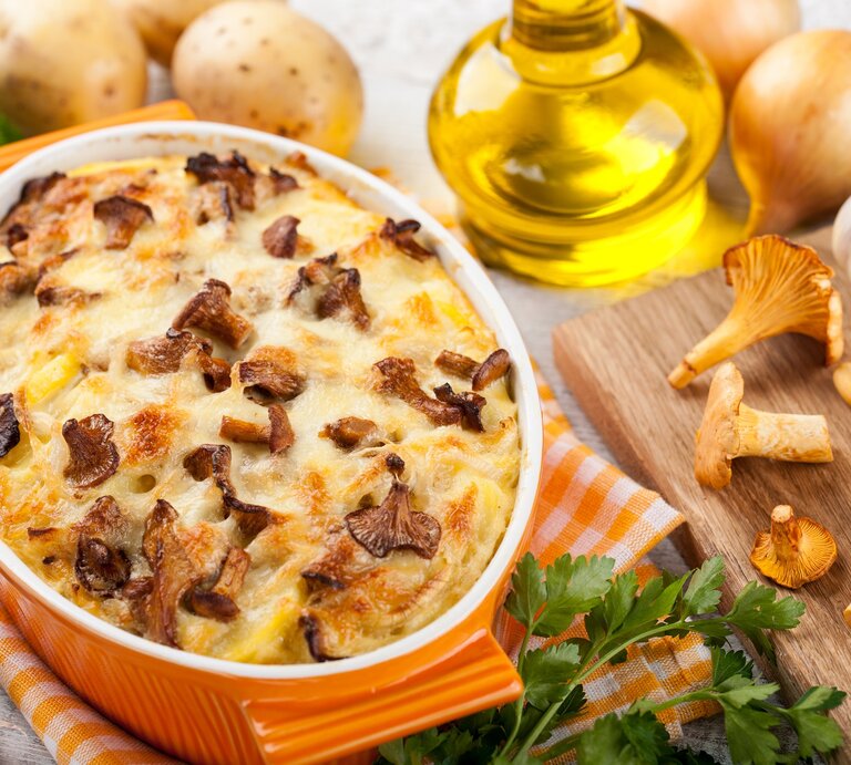 Kartoffelauflauf mit Pfifferlingen in einer gelben Auflaufform, drumherum frische Kartoffeln, Pfifferlinge und Knoblauch, außerdem eine Flasche Öl und Kräuter. | © Adobe Stock/ ffphoto