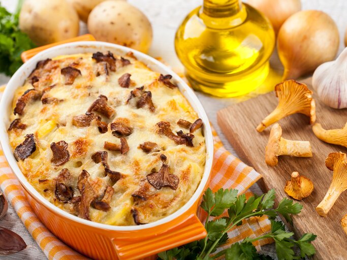 Kartoffelauflauf mit Pfifferlingen in einer gelben Auflaufform, drumherum frische Kartoffeln, Pfifferlinge und Knoblauch, außerdem eine Flasche Öl und Kräuter. | © Adobe Stock/ ffphoto