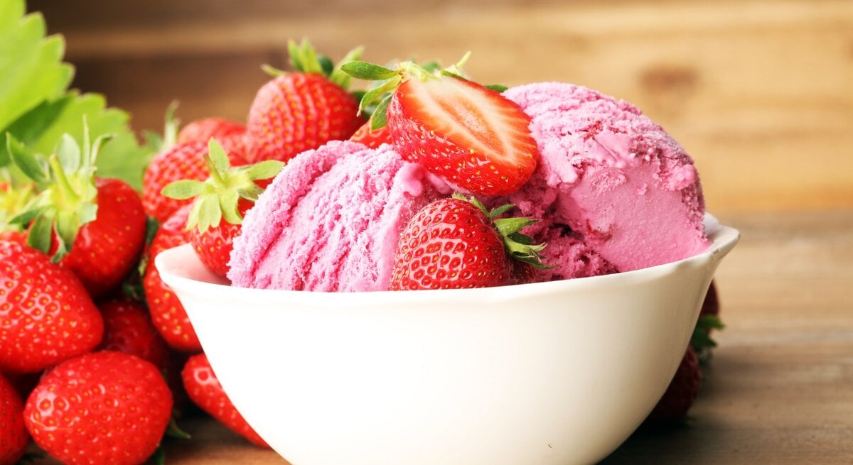 Frontal: eine weiße Porzellanschale mit zwei Kugeln Milchmädchen-Eis mit Erdbeeren auf einem Untergrund aus Holz.