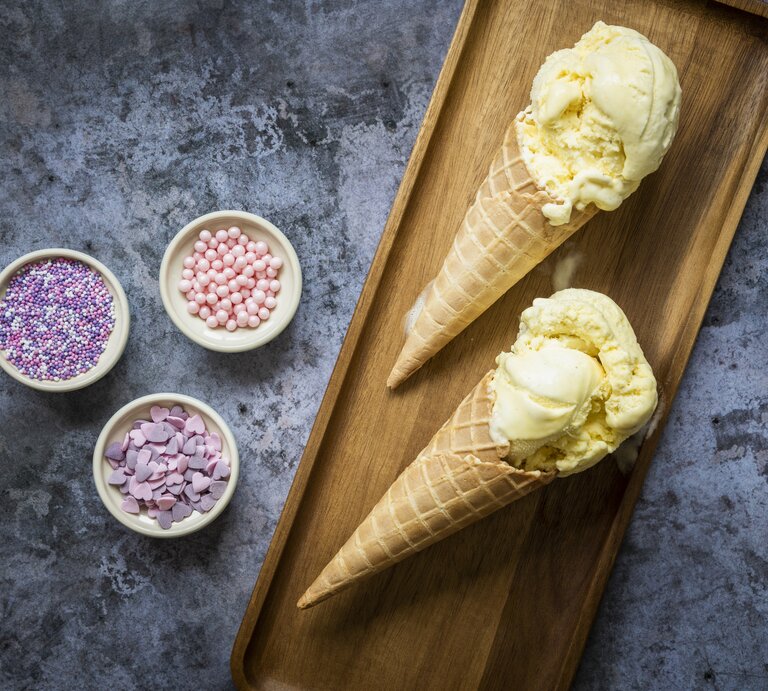 Eiscreme in Waffeln auf einem kleinen Holzbrett mit bunten Zuckerperlen auf einem dunklen Steintisch. | © GettyImages/ Anthia Cumming