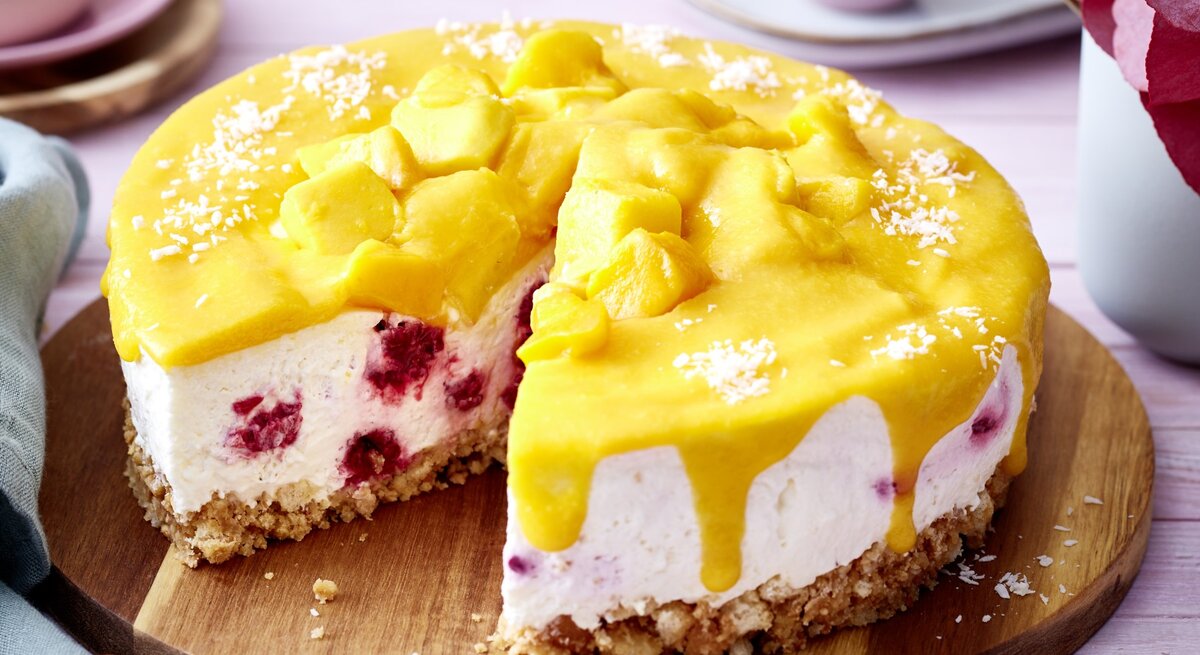 Angeschnittene Mango-Himbeer-Torte auf einem runden Holzbrett, heller Untergrund und helles Geschirr verschwommen im Hintergrund. 