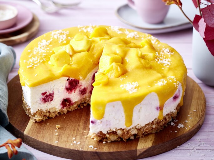 Angeschnittene Mango-Himbeer-Torte auf einem runden Holzbrett, heller Untergrund und helles Geschirr verschwommen im Hintergrund.  | © EAT CLUB / Isabell Triemer