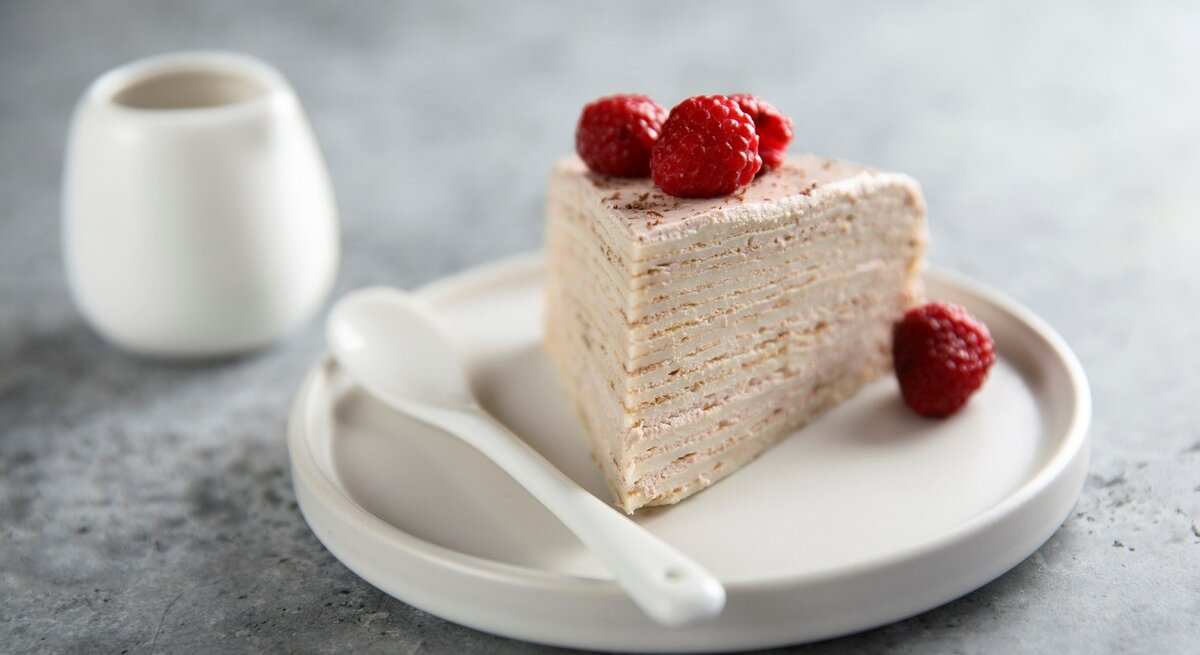 Auf einem Teller liegt ein Stück des Himbeer-Crepe-Kuchen. Man erkennt die Pfannkuchen-Schichten und die Cremefüllung. Er ist dekoriert mit Himbeeren.