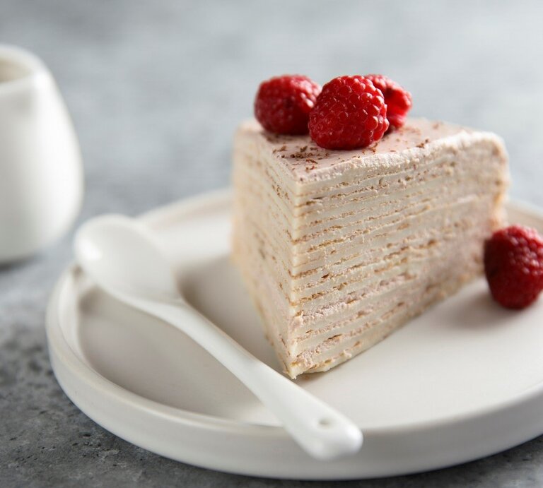 Auf einem Teller liegt ein Stück des Himbeer-Crepe-Kuchen. Man erkennt die Pfannkuchen-Schichten und die Cremefüllung. Er ist dekoriert mit Himbeeren. | © Getty Images / Mariha-kitchen 
