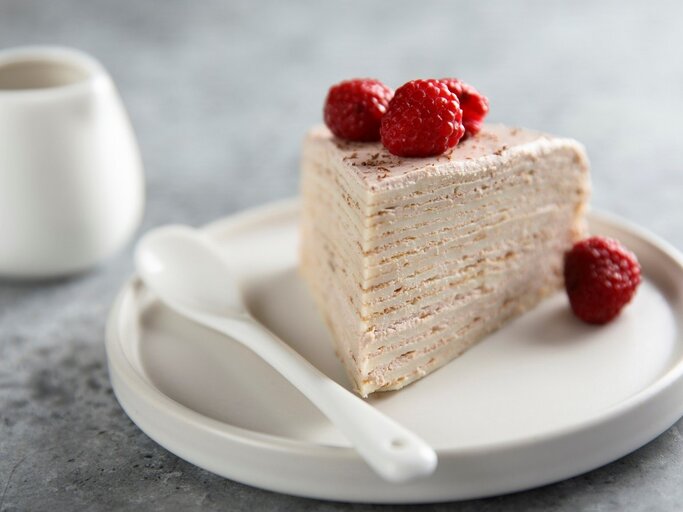 Auf einem Teller liegt ein Stück des Himbeer-Crepe-Kuchen. Man erkennt die Pfannkuchen-Schichten und die Cremefüllung. Er ist dekoriert mit Himbeeren. | © Getty Images / Mariha-kitchen 