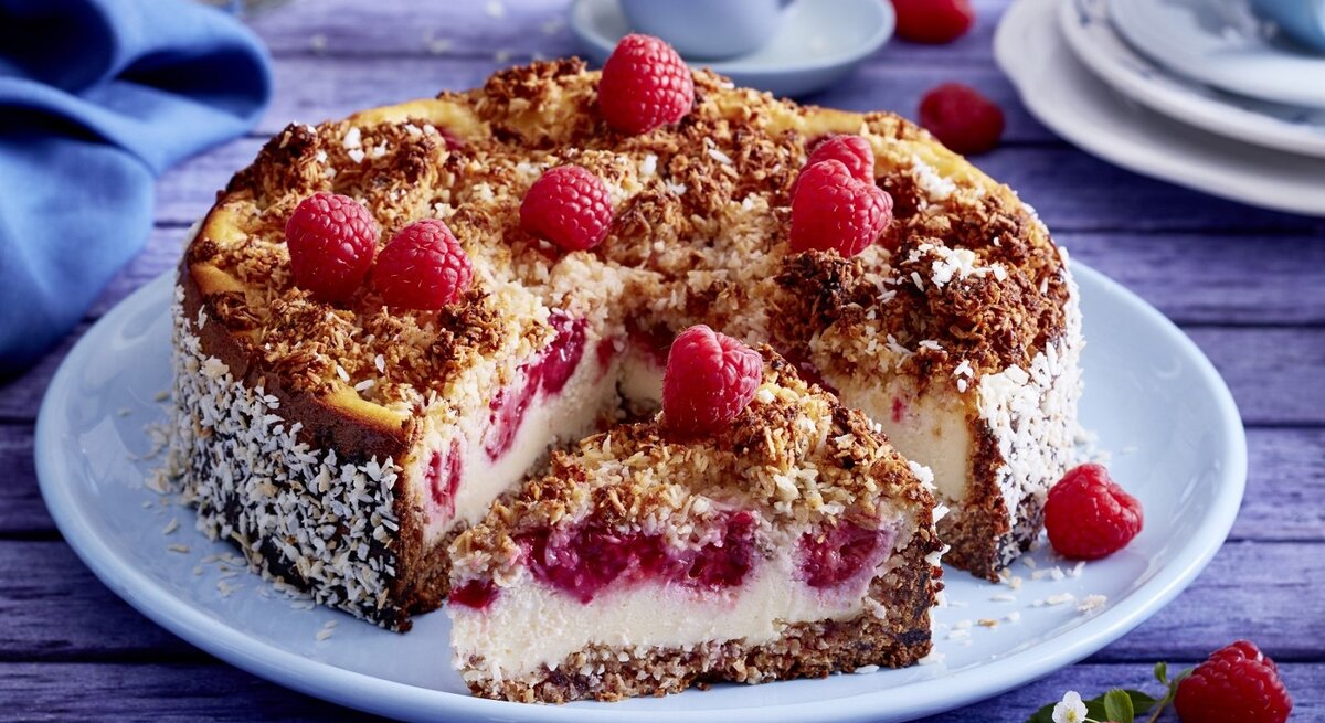 Auf einem Teller liegt ein Schmandkuchen mit Himbeeren. Er ist mit Himbeeren und Kokosraspeln dekoriert.