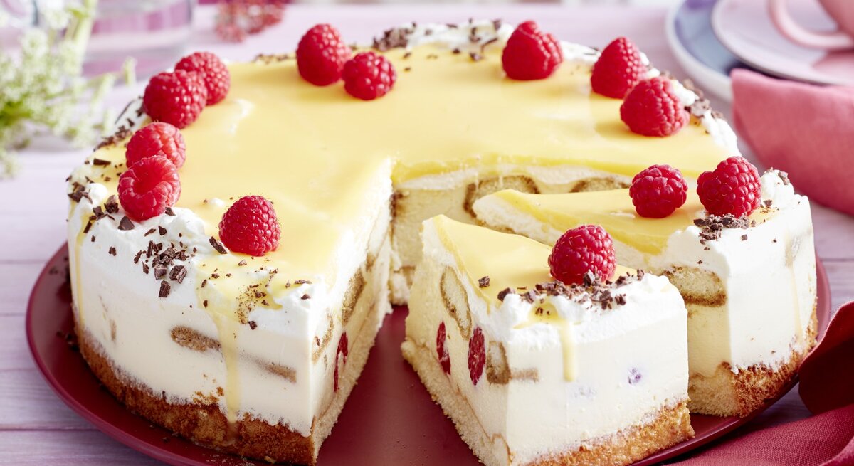 Auf einem Tortenteller steht eine Eierlikör-Himbeer-Tiramisutorte. Sie ist mit Himbeeren und Schokoraspeln dekoriert.