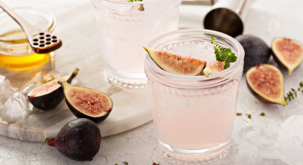Zwei Gläser Feigen-Spritz mit frischen Feigen und Kräutern, drumherum Bar-Zubehör und Zutaten.