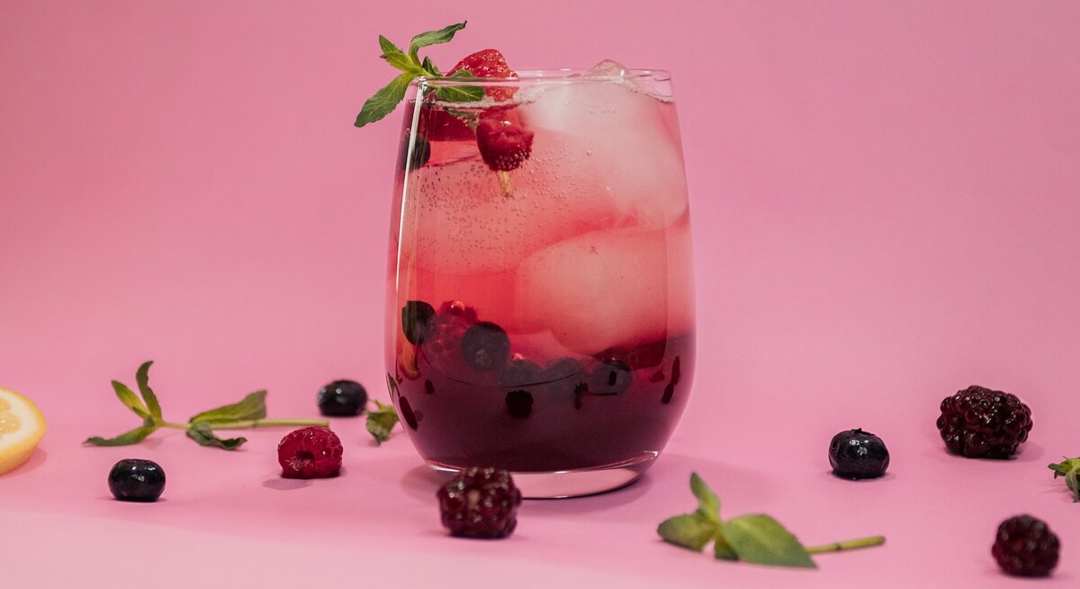 Ein Glas mit Lillet Spritz, Eiswürfeln, Beeren und Minze vor rosa Hintergrund, drumherum liegen Beeren und Minze.