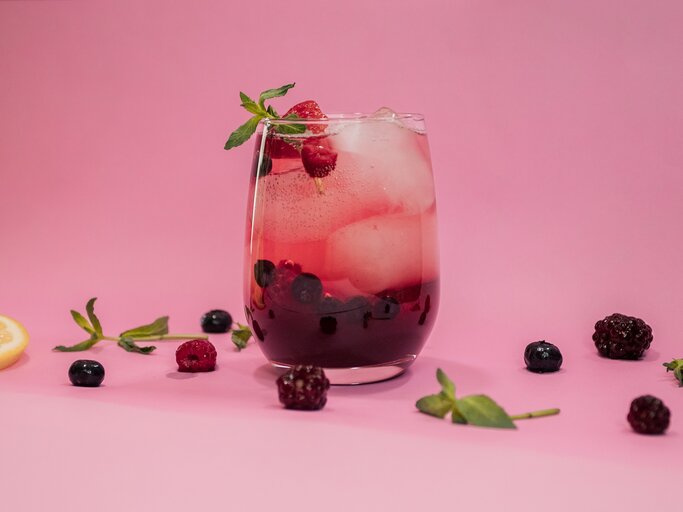 Ein Glas mit Lillet Spritz, Eiswürfeln, Beeren und Minze vor rosa Hintergrund, drumherum liegen Beeren und Minze. | © Adobe Stock/ francisco