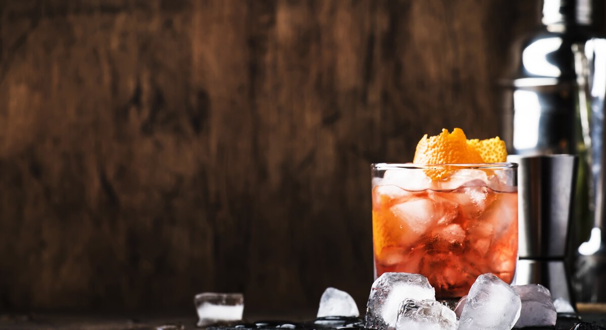 Ein Glas Aperol Sour und ein Cocktail-Shaker stehen in einem Raum aus Holz. Daneben liegen Eiswürfel. 