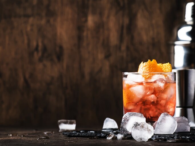 Ein Glas Aperol Sour und ein Cocktail-Shaker stehen in einem Raum aus Holz. Daneben liegen Eiswürfel.  | © Getty Images/5PH